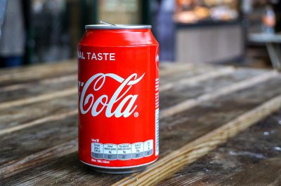 A photo of an unbranded Coca-Cola can sitting on top of a table, taken from above. The visible label includes some text about taste. It is placed in front of blurred out background elements like wooden tables or other items that could have been used for product photography. This setup would be perfect to capture product details and colors without any branding information being too prominent. The overall mood should convey a sense of simplicity and minimalism in the style of product photography.