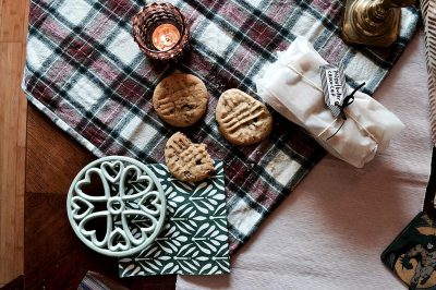 A plaid blanket on the floor with cookies and tea, wrapped gifts, on top of it is an intricately designed green and white patterned napkin with heart shaped designs, a small ceramic dish filled with chocolate chip cookies sits next to it, a lit candle rests in front of them, a gold ashtray stands beside them, a warm and cozy atmosphere fills our room, product photography, still life focus, shot from above, close up.