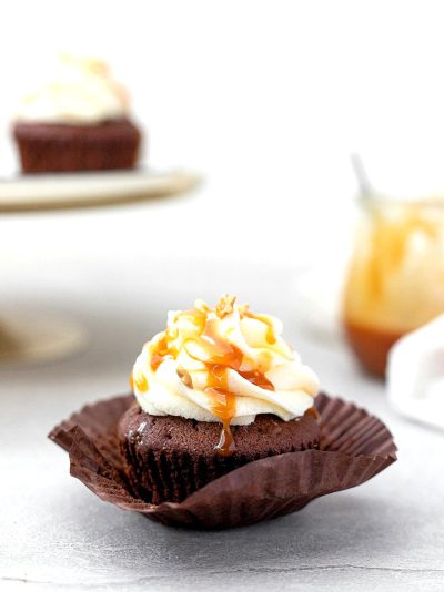 A photo of a rich, fudge brownie cupcake with caramel cream cheese frosting and drizzled caramel sauce on top. The background is white with two more cupcakes in the distance. The focus should be sharp to capture every detail of texture and color.