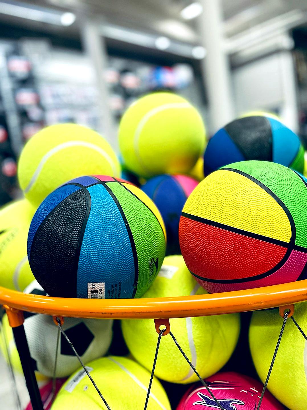 A basket of colorful tennis balls and basketballs, captured in closeup photography with a Canon EOS R5 camera. The background is an outdoor sports store. In the foreground, there is a yellow and black rugby ball placed on top of all other equipment. There should be no text or logos visible on any items.