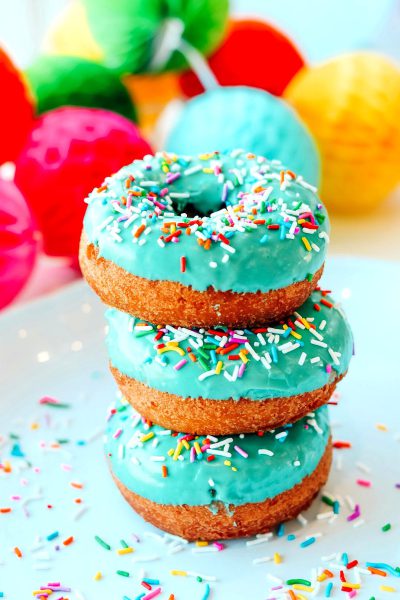 food photography of three stacked donuts with turquoise glaze and colorful sprinkles, bright background with party decorations in the back, vibrant colors, daylight