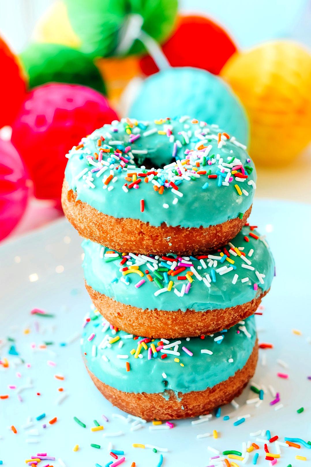 food photography of three stacked donuts with turquoise glaze and colorful sprinkles, bright background with party decorations in the back, vibrant colors, daylight