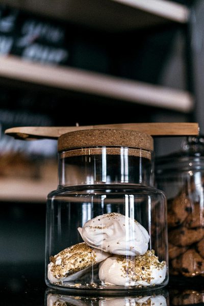 A glass jar with gold glitter meringue cookies inside, the lid is made of light brown cork and stands on top of it. The background features dark wooden shelves and a blurred black wall behind them. The kitchen table where all these elements stand also has an elegant white marble surface. The scene exudes elegance and sophistication. The product photography uses natural lighting with sharp details and professional color grading. The minimalistic style was captured with a Nikon D850 camera using a Nikon AFS Nikkor 70-200mm f/4G ED VR lens in a style reminiscent of professional photography.