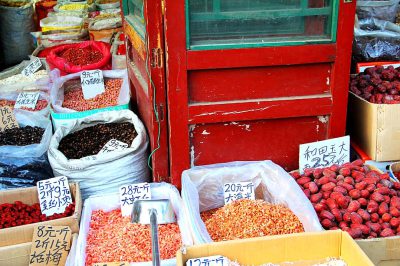 A red wooden stall in the Chinese market is filled with various dried fruits, such as dates and frozen strawberries, which have white signs displaying their price tags. The dried fruit bag on top of one bin has pink grabbers attached at its edges. In front of it sits an empty box containing red beans that appear fresh. There's also another small cardboard carton full of blackened winter sorghum beads, and several other boxes behind the stall selling different types of dried berries.