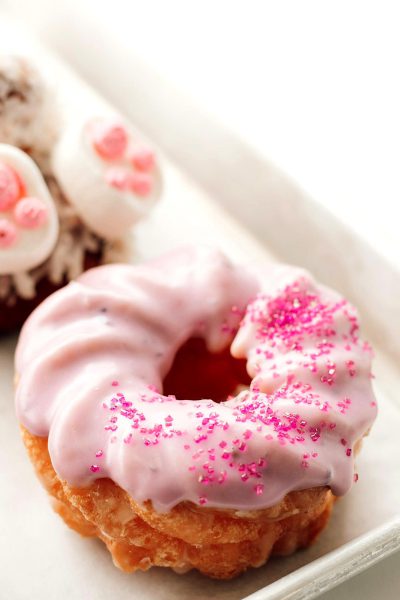 Close up photo of a pink donut with white glaze and little pink sprinkles, on the background there is another pastry in the style of the same, white plate, food photography, high key light