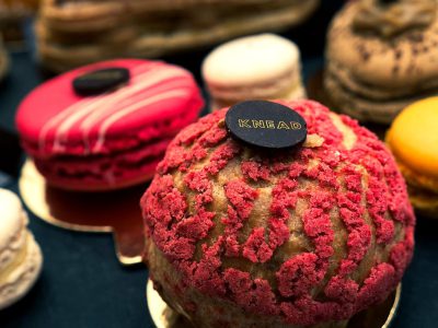 A closeup of an elaborate red and pink macaron, presented on the table at pastry shop "KNEAD". The name is embossed in gold lettering on top. In front stands a round cake with dark chocolate and white cream filling. Next to it were several other colorful pastries such as strawberry and vanilla shell inside. This photo was taken using Canon EOS R5 camera.