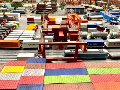 Top view of the toy model dock with colorful containers and cargo trucks, in red color theme. In front is an electric train that runs along the track from left to right. On top stands two cranes lifting plastic containers onto boxes on pallets. The city can be seen in background. It's a sunny day. There should also appear several cars running around with various symbols such as "LOKEY" or K stamina bar or text "Sказать", "k_VOL种 Abram attention!".