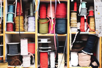 A closeup of various spools and rolls of different colors, sizes, textures, materials, shapes, and types of ribbon neatly arranged on shelves in the sewing studio's thread cabinet. The photo captures intricate details like ribbons with lace or braided edges, as well as unique patterns such as stripes, dots, hearts, stars, chevrons, etc., showcasing diversity within one material type.