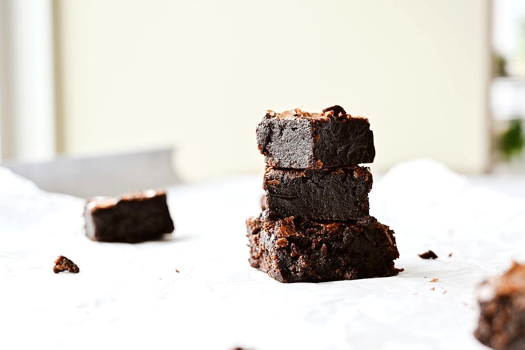 food photography of three fudge brownies stacked on top of each other, on white table cloth, with small pieces scattered around the base and a blurred background, taken from an angle