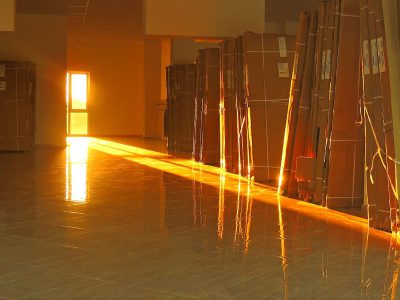 In the early morning, sunlight shines on several large cardboard boxes placed in an empty room with light brown floors and walls. The warm orange glow reflects off them as if they were made of glass or metal. In front is a long beam that adds to their shine. There's no furniture visible but some small items scattered around, adding depth to the scene. This photograph captures the beauty of light shining through objects.