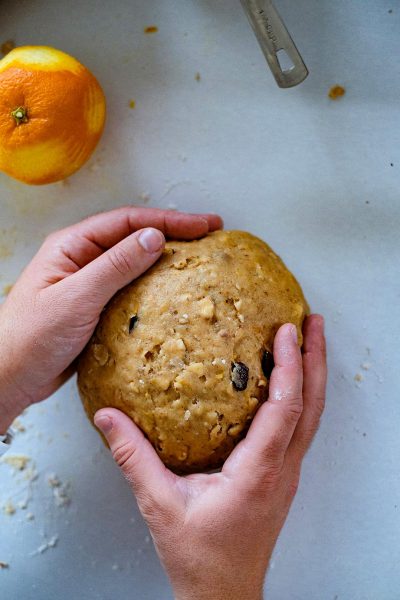 a person's hands holding and kneading an orange sweet potato bread dough, on white kitchen counter with the edge of the bowl for sweets in it. In background is half oranges and one dried fruit or chocolate chip inside, and a few flour dust around. top view, aerial shot, captured by Hasselblad X2D camera.