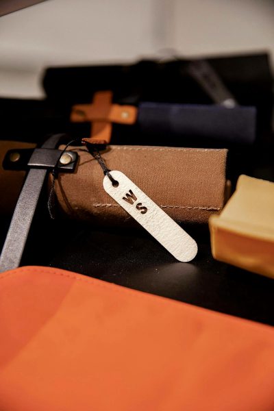 close up photo of leather tag with text "B", on top of black table, next to orange bag and navy blue small case made from wool felt fabric. There is also some tools in the background. The lighting is soft and natural. Shot by [Tim Walker](https://goo.gl/search?artist%20Tim%20Walker)