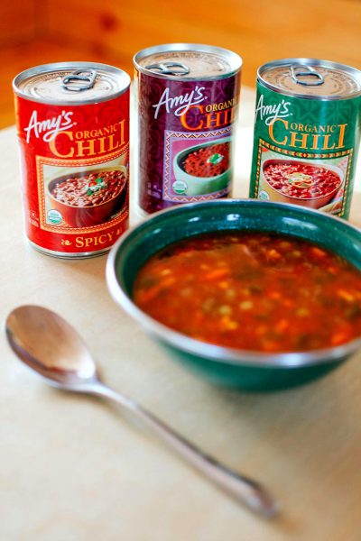 A photo of three cans of "amys organic chili" on the table with one can open and a bowl filled half way with soup, spoon next to it. The top two cans have colorful labels showing different types of chilis. Bright natural light illuminates them from above. A warm feeling is created in the style of this scene.