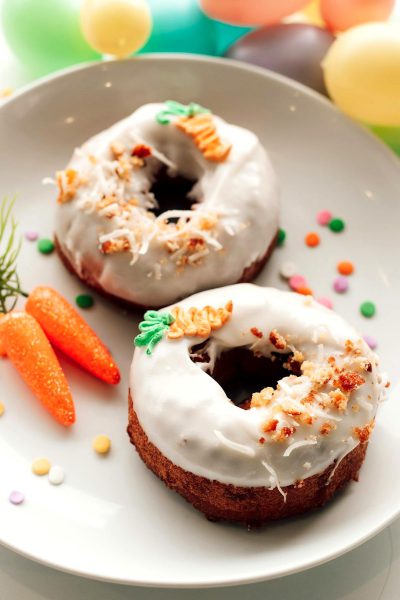 Easter-themed air-fried carrot cake donuts, decorated with coconut shavings and carom dots on top of the glaze, served on an elegant white plate, with Easter eggs and carrots visible in the background in a close-up shot with natural light.