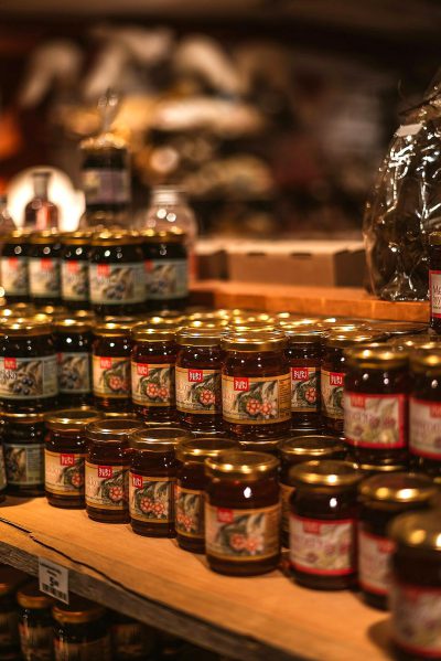 A closeup shot of jars and bottles filled with various types of jam, arranged on shelves in the background of an Italian fish shop, creates a contrast between the jar product display and traditional fishing elements. The focus should be sharp to capture details like labels or branding on each jelly pot, highlighting their diversity and quality, with warm lighting that highlights textures and colors, creating depth in the scene.