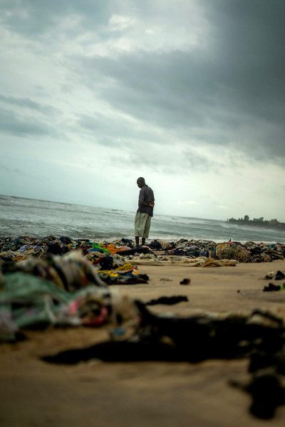 A photograph of an African man standing on the beach, looking at large piles of garbage and  scattered across the shore. The ocean is visible in the background with stormy clouds overhead. A sense of hopelessness hangs over everything as he looks out to sea.