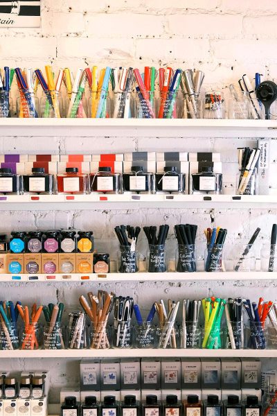 A photo of shelves filled with art supplies like markers, ink and watercolor paints. The focus is on the colorful tubes of markers that stand out against the white brick walls. There is an array of other artistic tools such as paintbrushes, scissors and various vials for inks. This setting creates a creative environment where artists can take inspiration from these materials to create their masterpieces in the style of various artists.