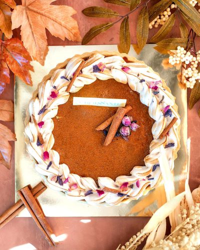 A topdown view of an autumn pie, adorned with cinnamon sticks and rose petals in the center. The sides have braided cream cheese frosting patterns resembling rope or ribbon. A white label is placed on one side of it. In front of the cake lies two c.FileOutputStreams. On the right corner there's dried leaves and wildflowers.