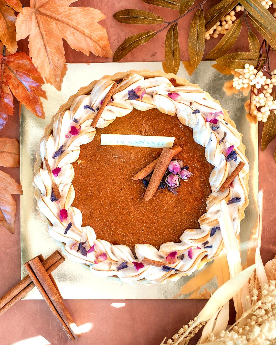 A topdown view of an autumn pie, adorned with cinnamon sticks and rose petals in the center. The sides have braided cream cheese frosting patterns resembling rope or ribbon. A white label is placed on one side of it. In front of the cake lies two c.FileOutputStreams. On the right corner there’s dried leaves and wildflowers.