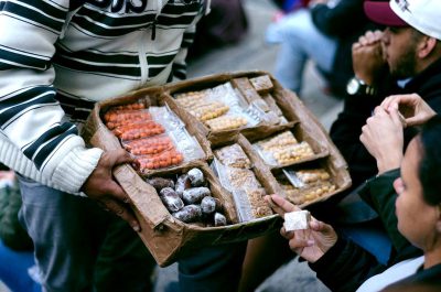 A photo shows people selling food in the streets, holding boxes with various snacks inside like nuts and dried fruits, surrounded by other passersby with their hands open to take them. The composition looks down from above and features a closeup shot.