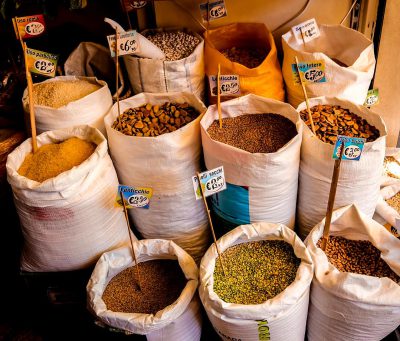 Sense of color, stock photo of large white bags filled with various types and sizes of grains at a market stall in a Spanish town center, next to blue signs with price tags showing $2 per bag. In one part is cumin powder, in another green herbs, dried beans. The background shows an old wooden table top with some more scattered grain. Bright warm light from inside. The style evokes a realistic painting in the style of an Old Master.