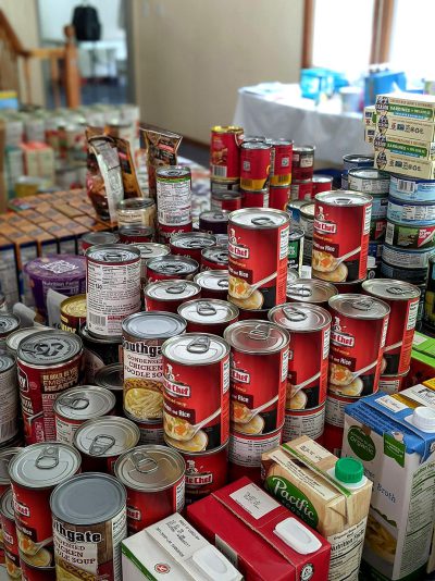 A pile of cans, boxes and packaging from various food brands were on the table at home for an important community Food drive to help those in need. The feeling showed people coming together with love to help others who need it most. There was a sense of unity as well as hope that we can all make a positive impact on our world by having this type of event. People stood ready to do work so they will be there for the local community. The cans showed different food brands without text or logos.