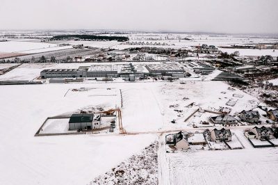 Aerial view of the city's industrial park in winter, surrounded by white snow and farmhouses on both sides. The warehouse complex is located at one end of an open field with houses nearby. In the style of Scandinavian photography, it features high-definition images with a gray sky, wide-angle lenses, distant views, pure colors, and static composition.