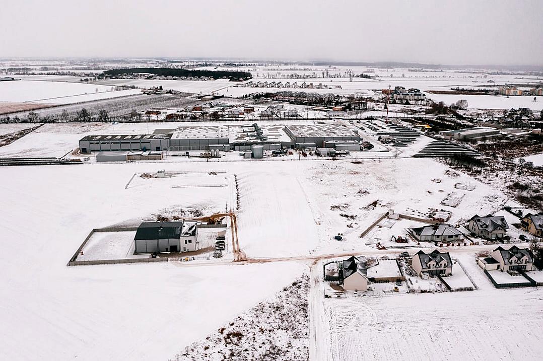 Aerial view of the city’s industrial park in winter, surrounded by white snow and farmhouses on both sides. The warehouse complex is located at one end of an open field with houses nearby. In the style of Scandinavian photography, it features high-definition images with a gray sky, wide-angle lenses, distant views, pure colors, and static composition.