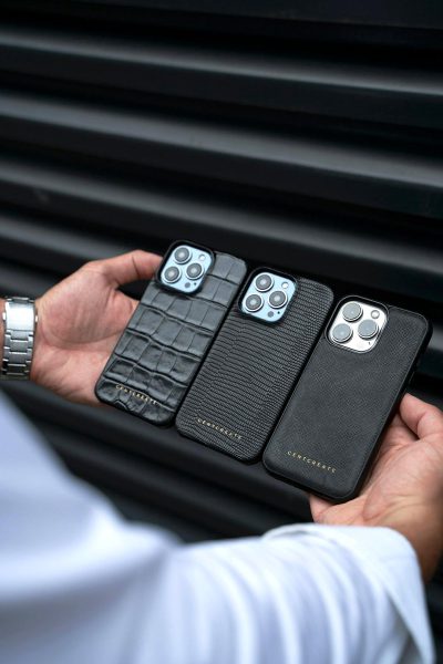 A hand holding three black leather phone cases for the iPhone, each with different textures and colors. The first case is made of crocodile skin material, the second from snake texture, while the third has a smooth surface in gray color. Each cover bears an embossed logo on its side that reads "CCroweggate". In front of them stands a man wearing white sleeves and pants, his face not visible due to being turned sideways towards us. He holds all the phones in the style of their ha resting against a car door.