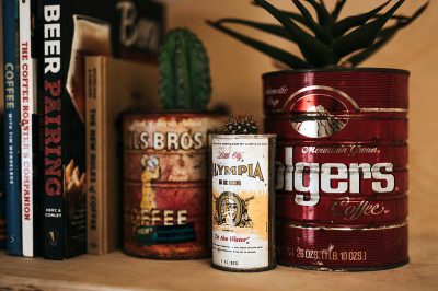 A vintage-inspired coffee can with "Olympia" and the phrase 'shy bros', striking colors of reds and golds, on display next to books that say things like beer cans, cacti, and sports cards. The scene is set in an old-fashioned home office or bar corner in the style of vintage product photography.