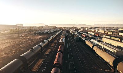 A photo of an industrial train yard in Arizona taken from above with the sun shining on the ground. There is a single freight box car and several oil tank cars on tracks, the sky has a few clouds and it is late afternoon, shot in the style of Hasselblad X2D, cinematic still, cinematic light, hyper realistic.