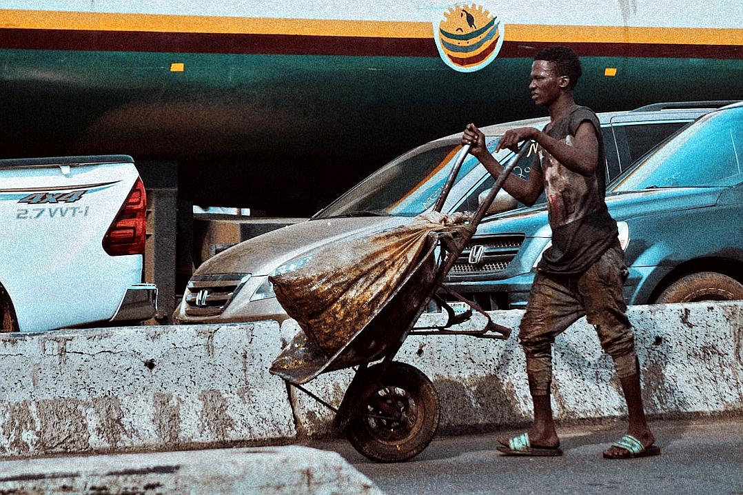 A photo of an African street kid pushing a wheelbarrow full of garbage. He is wearing worn out shoes and is barefoot. In the background, there is a blue SUV parked on the concrete border next to a motorway highway bridge with a happy smiley sign. The photo is in the style of Anthropic.