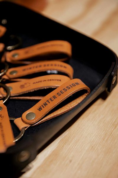 Photograph of leather key fob designs with the words "Winter session made in denver" engraved on them, laid out inside an open black case that is sitting atop a light wood table top. The background should be simple and neutral to keep the focus on the product.