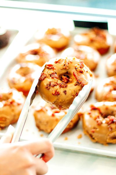 A close-up photo of someone taking a maplebourbon praline donut with bacon from the tray, there are other doughnuts on it against a white background.