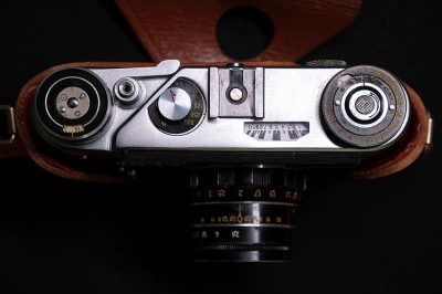 A close up of the top part of an old camera with a silver body and leather case, showing its focus circles on one side, its Leica emblem on another, its front view on the right side, its F08 setting in black ink text and its f2 lens, all set against a dark background. The photo, taken from above, focuses only on these details without any other surrounding elements. The photo is in the style of a minimalist aesthetic.