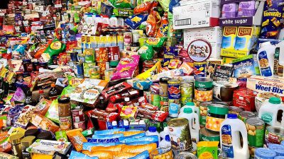 A large pile of food and goods, including several boxes with "white sugar", many bags of chips, some cans of used beans, various smoothies in plastic bottles, colorful red boxes filled to the brim full of cookies, etc., with the entire scene captured from above. The background is a grocery store with other items such as chocolate bars, milk cartons, white rice and more. It's a bright day outside, with sunlight casting shadows on all objects, creating an atmosphere of abundance and fast characteristic life style.