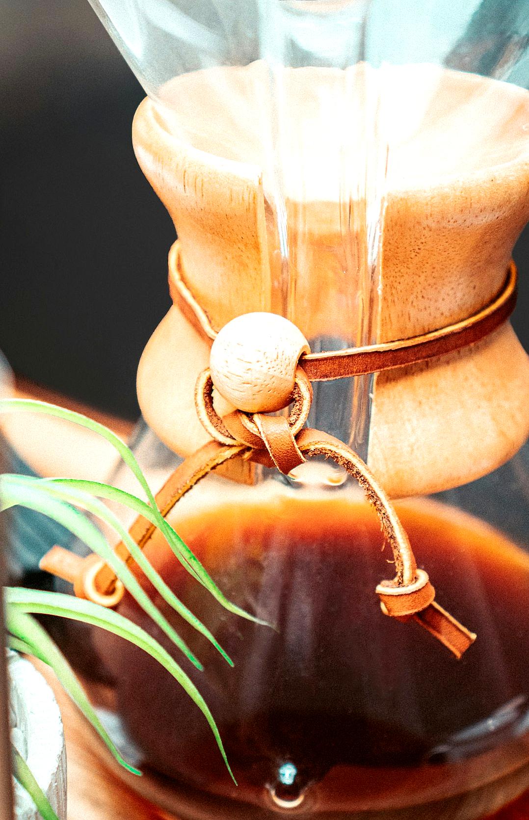 A closeup of the leather necktie on a coffee dripper in the style of Art osaka, with rich brown hues and subtle stitching details visible against a backdrop of swirling coffee in a glass carafe. The background is softly blurred to focus attention on the detailed texture of the leather. A small green plant adds contrast and color to the scene.