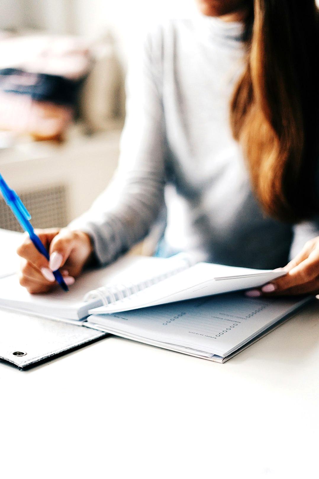 A woman writing in her notebook, sitting at a table with an open notepad and pen on it, against a white background, in a closeup shot of hands using a blue ballpoint pen to write notes, with soft focus, natural light from a window, in a modern interior design. The atmosphere is calm and focused, highlighting attention, in the style of modern interior design.