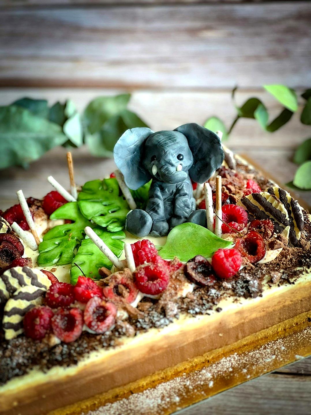 A rectangular chocolate and cream tart with an elephant motif, surrounded by teal leaves and raspberries, with black sweets on top and candles around it. A grey baby elephant figurine is placed in front of them. A soft background with eucalyptus branches. A realistic photo in the style of nature photography.