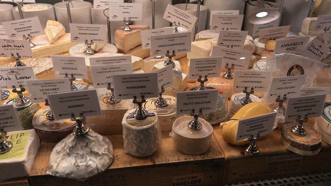 A photo shows a cheese display with white labels on each piece, with each label having the name and description of the cheese in French or English. The wooden counter has several neatly arranged types of cheese. In front is an empty space for customers to place their selections. On top are three silver stands holding small signs also written in French. On one side you can see other craft supplies such as twine, cream-colored boxes, and so on.