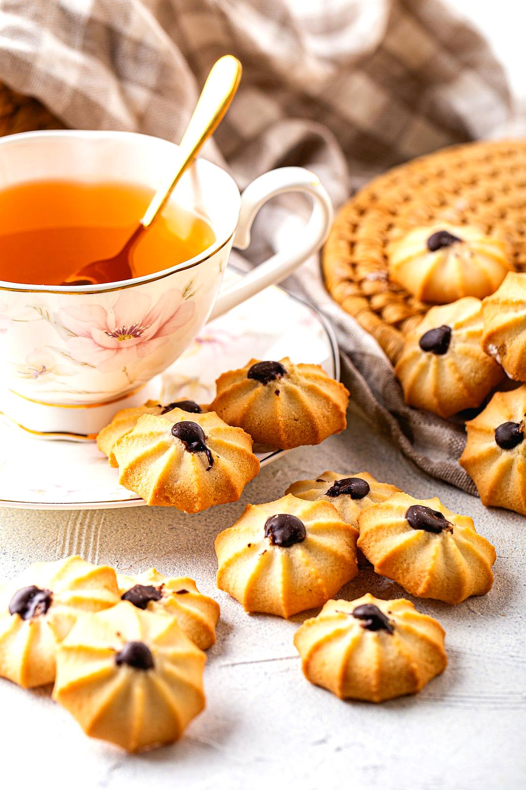 A photo of delicious-looking freshly baked cookies with chocolate chips and some cut into flower shapes, placed on the table next to an elegant teacup filled with tea. The background is a light gray or white, creating a soft and inviting atmosphere. A kitchen towel hangs in the back for serving. The photo is in the style of a minimalist artist.