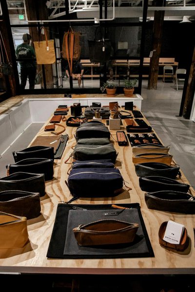 A long table with various leather pouches and bag styles was displayed on it, showcasing the variety of designs in the style of The accoutrements company in an industrial space. In front was empty black paper for customer iphone photos. It was a product display presentation set up for people to walk around. A person dressed as a crew member stood next to one side of the tables.