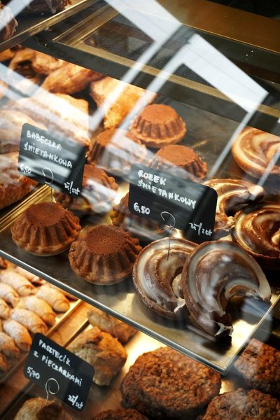 A closeup of the display case in an urban bakery, showcasing various pastries such as cakes and cookies with price tags. The glass panel reflects light from above, creating a soft glow on each item. A black sign shows that one pair is "6!", while another says "3ernelik." This scene captures focus to highlight details like intricate patterns or textures on different types of delicious treats in the style of impressionism.