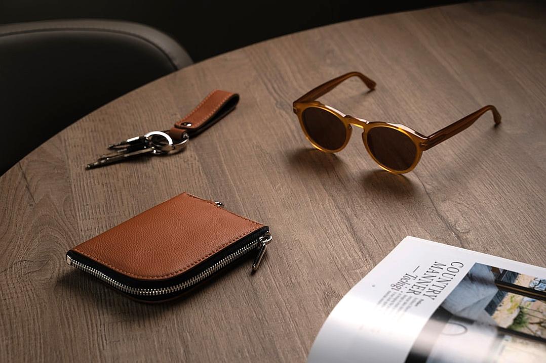 A leather brown wallet with the zipper open, sunglasses and car keys on top of it, a magazine spread next to that, placed on an oak table in a minimalist style with soft lighting for product photography.