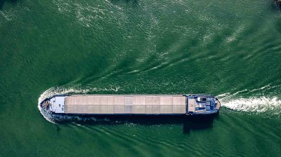 Aerial view of a barge in a green river, with white waves and ripples behind the ship, with motion blur, taken with drone photography, with a flat perspective, in the style of minimalism, from a top down view, of the water surface, during daylight.