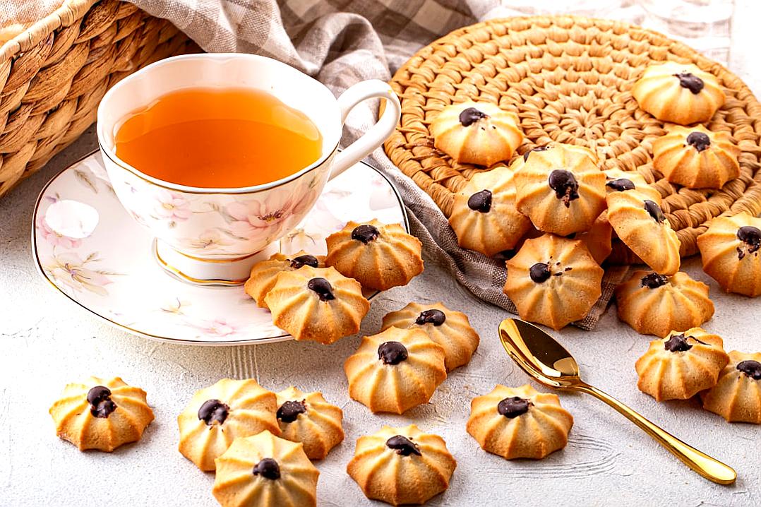 A photo of mini cookies with chocolate chips in the shape of flowers on top, placed next to an elegant teacup and saucer filled with tea. A wicker basket is also visible nearby, and there is some fabric around it all. The background features light grey concrete and white linen cloth. This scene has warm tones, and golden cutlery adds sophistication. The photo has the style of a minimalist painter.