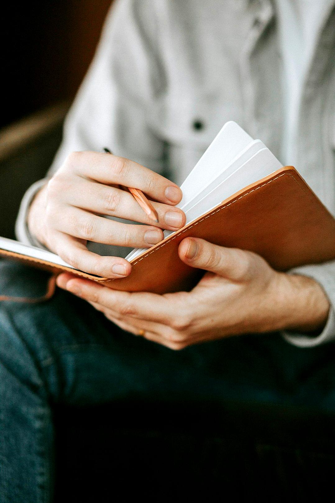 A closeup shot shows hands holding an open leather notebook. One hand turns the pages as the other hand writes on it. The background is blurred to focus solely on the fingers turning the page in a detailed manner. He is wearing casual attire and sitting comfortably inside, embodying a moment of self reflection or creation. The style resembles that of an artist.