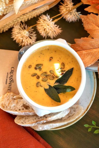 A white bowl of creamy butternut pumpkin soup with scattered seeds and garnished in the style of sage leaves, on top of sliced baguette bread, placed next to an autumn-themed card reading "LA CRUSSOLSYN TRANStOTE", all set against the backdrop of fall foliage. The scene is illuminated by soft natural light, creating a warm atmosphere that suggests comfort and hominess. High resolution photography, high definition, hyper realistic.