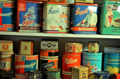 A collection of vintage tin cans from the mid-20th century, displaying colorful labels and illustrations on their sides, arranged in an aesthetically pleasing display. The tins include "Villa Velliana" and other brand logos. They stand against a white wall with shelves beneath them filled to about one third full with similar antique items like coffee jars and candy boxes in the style of vintage illustrations.