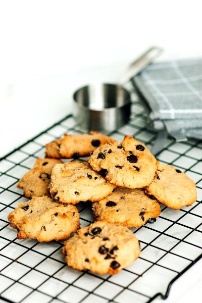 A photo of low downwards angle of small, round chocolate chip cookies on an iron wire rack with some kitchen utensils in the background. The white table top is visible and has soft lighting. A few blue napkins can be seen in one corner. There's no natural light source in sight, only artificial lighting that gives everything a warm glow. This scene creates a cozy atmosphere where you could feel like enjoying these sweet cookies as part of your breakfast or midm.Flowers on top of them, to make it look more real